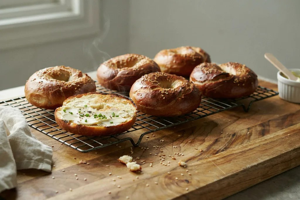 Sourdough bagels on a wire rack, deeply burnished from the boil, one split and toasted with cream cheese.