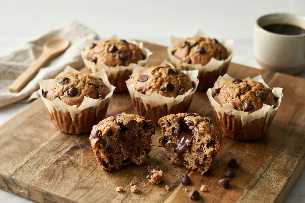 Whole grain chocolate chip muffins with craggy tops, one broken open showing molten chocolate through a speckled crumb.